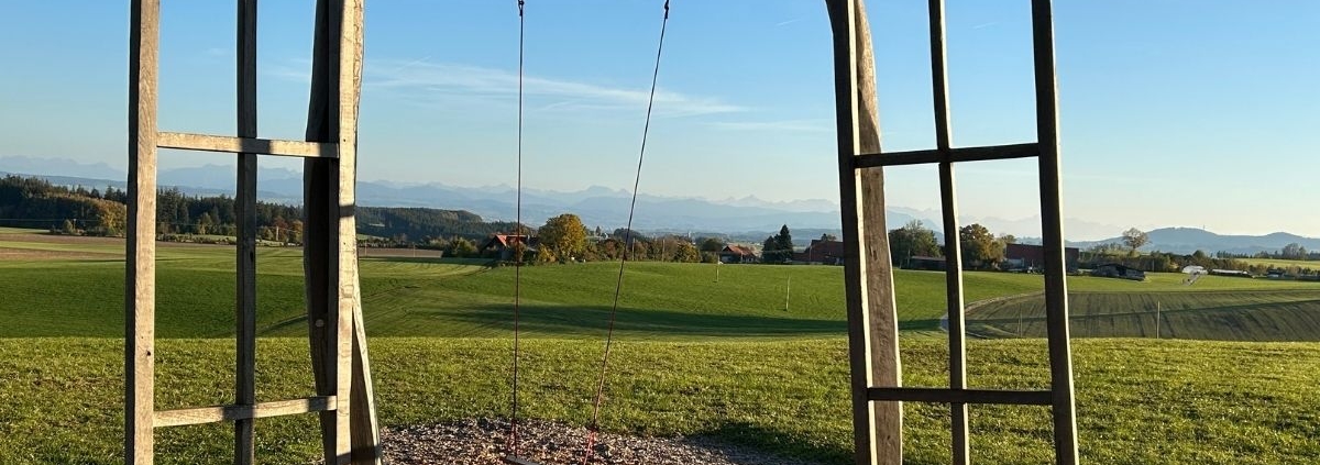 Offene Holztüren an einem freistehenden Rahmen mit Schaukeln, dahinter eine weite grüne Landschaft.