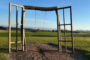 Offene Holztüren an einem freistehenden Rahmen mit Schaukeln, dahinter eine weite grüne Landschaft.